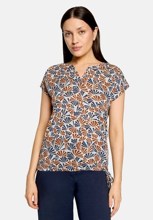Frau trägt eine kurzärmelige Bluse mit einem braunen, marineblauen und weißen Fächer-Muster und marineblaue Hose, steht vor einem einfarbigen Hintergrund.