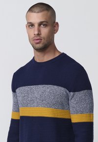 Navyblauer Pullover mit strukturierter Stricktechnik, der graue und gelbe horizontale Streifen aufweist, die ein geschichtetes Design erzeugen. Rundhals.