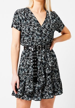 Kvinde iført en sort kort kjole med blomstermønster og bredt sort bælte med flere sølv-øjenhuller, poserer med den ene hånd på hoften.