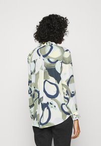 Una donna con capelli ricci indossa una camicia a fantasia con motivi circolari in tonalità di verde e blu, in piedi contro uno sfondo bianco semplice.