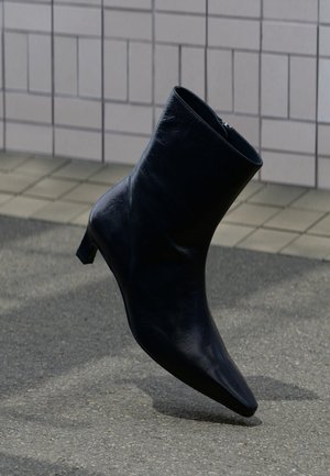 Stivaletto alla caviglia in pelle nera con tacco basso, inclinato su una superficie lastricata vicino a una parete piastrellata sullo sfondo.