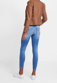 Bruine leren jas met gewatteerde accenten, gecombineerd met blauwe skinny jeans met een vervaagde wassing. Witte sneakers maken de outfit compleet.