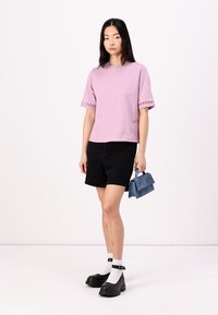 Lys lilla bomulls t-skjorte med korte ermer og øylet trim, kombinert med svarte shorts, hvite sokker og kraftige svarte sko. Holder en blå veske.