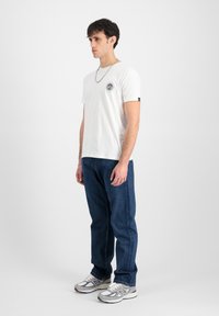 Hvid bomulds t-shirt med rund hals og et lille broderet logo på venstre bryst. Båret med mørkeblå jeans og grå sneakers.