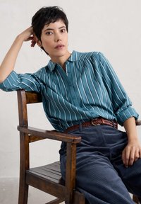 Bluzka w paski w odcieniu niebiesko-zielonym z kołnierzykiem, w zestawie z granatowymi spodniami z korduroju i plecionym brązowym paskiem, siedząca na drewnianym krześle.