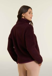 Donna con capelli scuri che indossa una giacca di pile borgogna e pantaloni beige, in piedi di lato con le mani in tasca su uno sfondo chiaro.