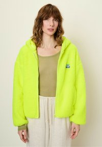 Neon yellow fleece jacket with a zipper and hood, featuring ribbed cuffs and a small logo patch. Worn over a light olive top and light pants.