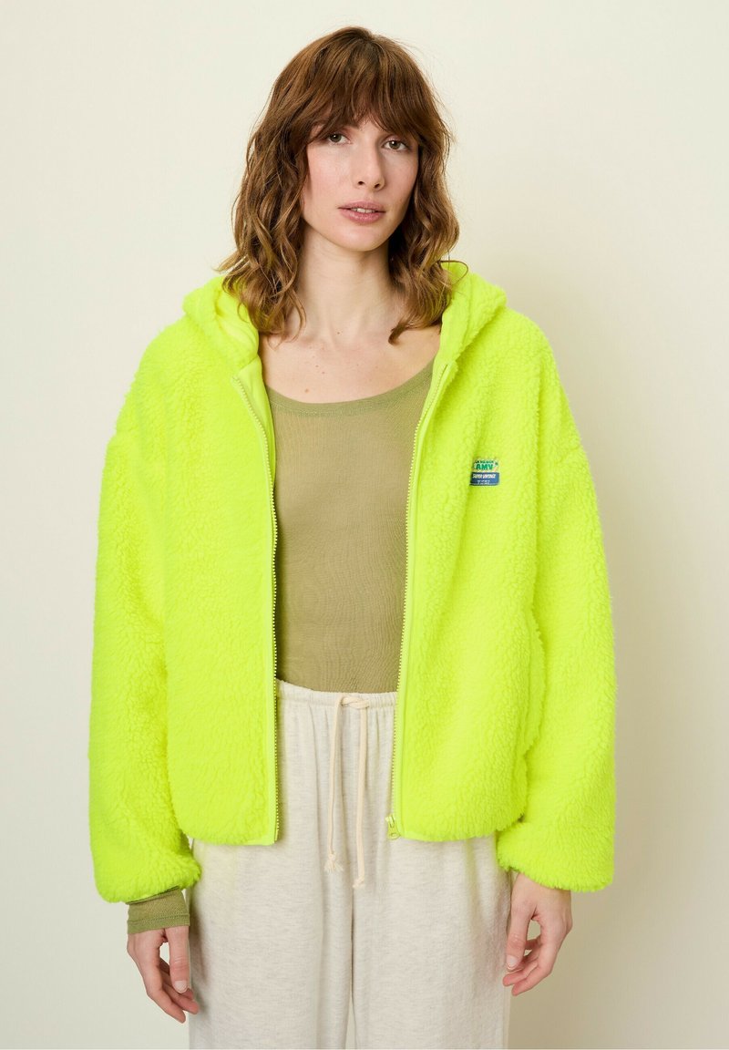 Neon yellow fleece jacket with a zipper and hood, featuring ribbed cuffs and a small logo patch. Worn over a light olive top and light pants.