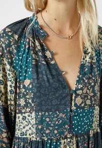 Robe fluide avec un design en patchwork incluant des blocs teal, floraux et à motifs. Encolure en V avec des accents de boutons et un détail de col à volants.