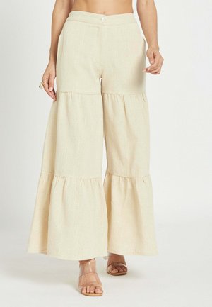 Pantalones beige de pierna ancha y de varias capas con cintura de botón y cremallera, combinados con tacones abiertos con tiras transparentes, mostrados de la cintura a los pies.