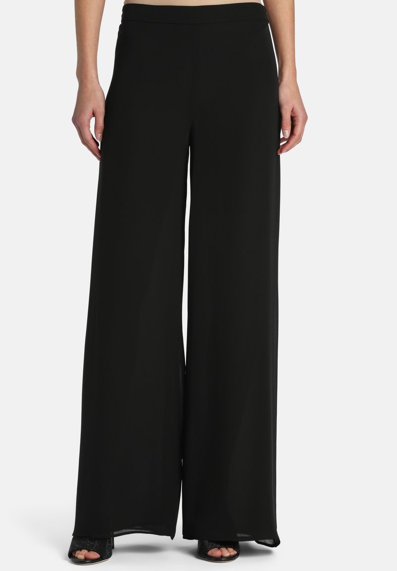 Personne portant un pantalon noir ample à taille haute avec jambes larges et des talons noirs ouverts, debout devant un fond clair uni.