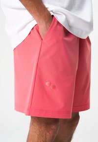 Wasserabweisende rosa Shorts mit einer glatten Textur, ausgestattet mit Seitentaschen und einem dezenten Logo-Detail am Saum. Leichtes und atmungsaktives Material.