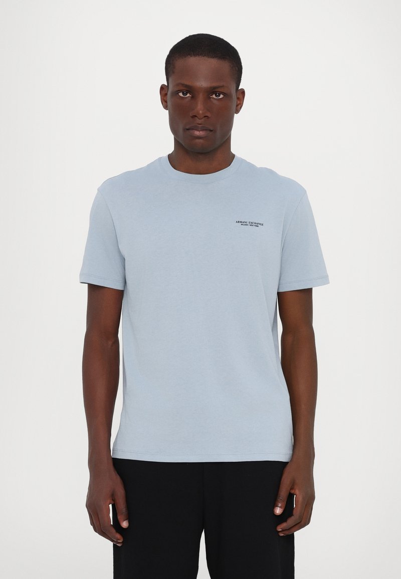 Gaiši zils kokvilnas T-krekls ar apaļu kakla izgriezumu un īsām piedurknēm. Tam ir neliels melns logotipa prints augšējā kreisajā krūtīs.