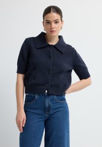 Marineblauer gestrickter Cardigan mit kurzen Ärmeln, breitem Kragen, fünf Knöpfen auf der Vorderseite und geripptem Saum, getragen über hochtaillierten blauen Jeans.