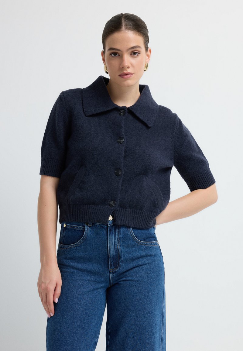 Marineblauer gestrickter Cardigan mit kurzen Ärmeln, breitem Kragen, fünf Knöpfen auf der Vorderseite und geripptem Saum, getragen über hochtaillierten blauen Jeans.