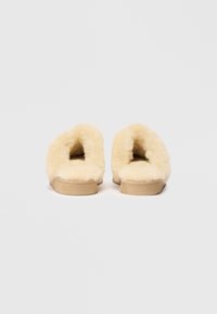 Pelsko med myk, fluffy beige fôr og en lys tan såle, med åpen bakdesign for enkel påkledning.