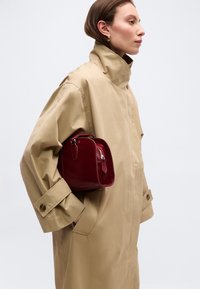 Γυναίκα με μπεζ καπαρντίνα με μεγάλο γιακά, κρατώντας μια μικρή κόκκινη δερμάτινη τσάντα σε απλό ανοιχτόχρωμο φόντο.