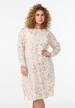 Kobieta o kręconych włosach, ubrana w jasną sukienkę o długości do kolan z długim rękawem i małym kwiatowym wzorem, stojąca z jedną ręką na biodrze.