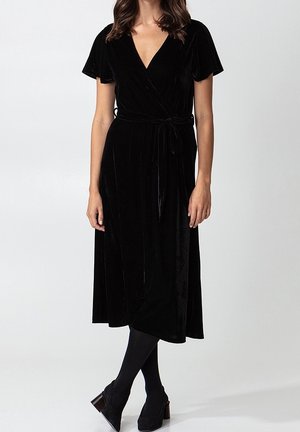 Femme portant une robe portefeuille en velours noir à manches courtes, associée à des collants noirs et des chaussures à talons blocs noires, debout sur fond blanc.