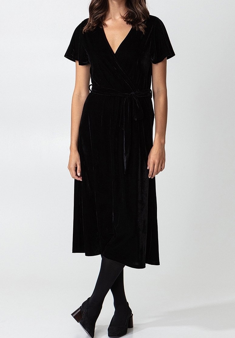 Femme portant une robe portefeuille en velours noir à manches courtes, associée à des collants noirs et des chaussures à talons blocs noires, debout sur fond blanc.