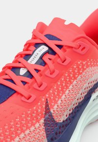 Běžecké boty Nike Pegasus v jasně růžové a námořnické modři. Vlastnosti zahrnují texturovaný síťovaný svršek, živé tkaničky a polstrovanou podrážku s detailem značky.