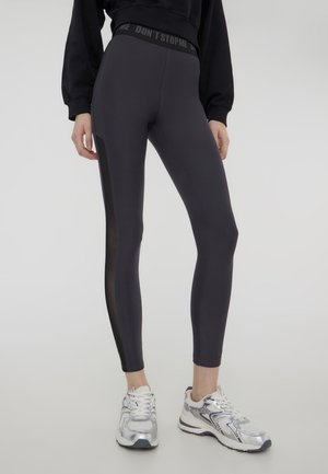 Leggings grigi scuri con una vita alta, caratterizzati da un pannello in rete nera sui lati e una texture liscia e elasticizzata.