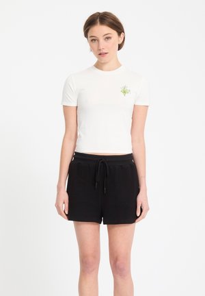 Jeune femme debout, portant un t-shirt blanc court avec un petit motif vert et un short noir à cordon, sur un fond blanc.