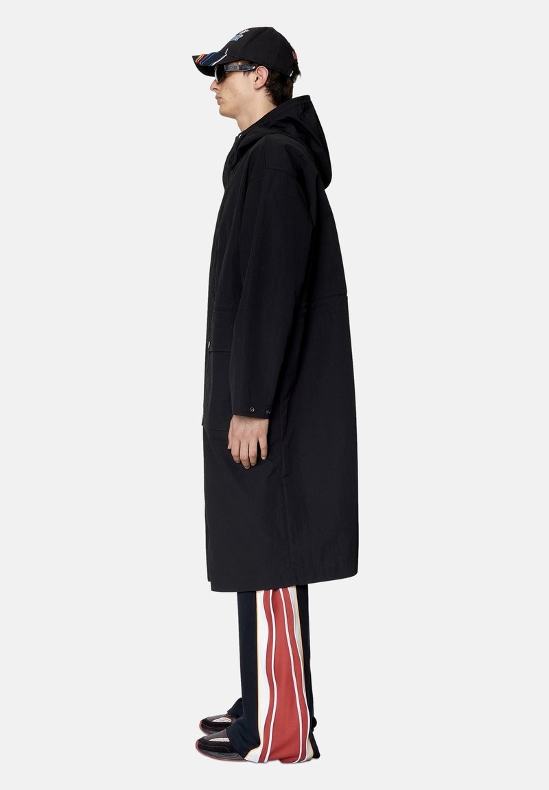 Jonge man in een lange zwarte hooded jas, zwarte pet, zonnebril, gestreepte wijde broek en zwarte schoenen, staande in profiel tegen een witte achtergrond.