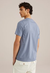 Světle modré tričko s krátkým rukávem a kulatým výstřihem, oblečené obráceně modelem, v kombinaci s krémovými kalhotami s volným střihem.