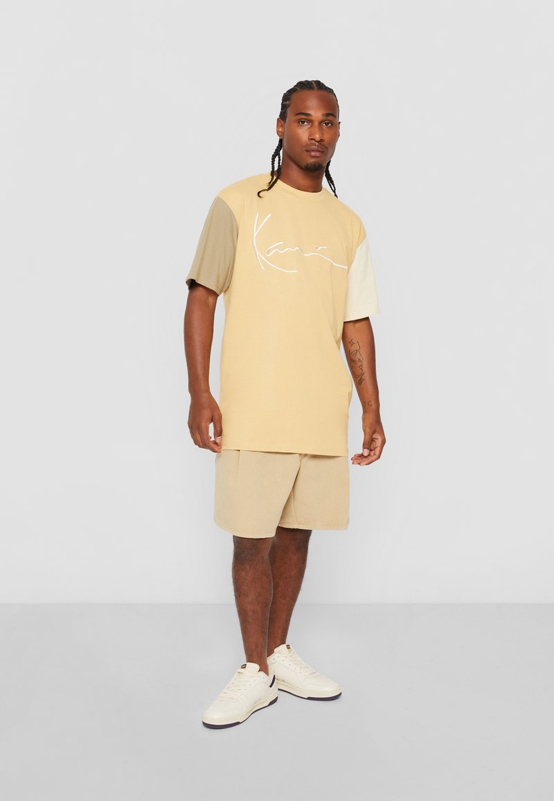 T-shirt à manches courtes en coloris beige et marron clair, arborant un logo en script blanc. Porté avec un short beige assorti et des baskets blanches.