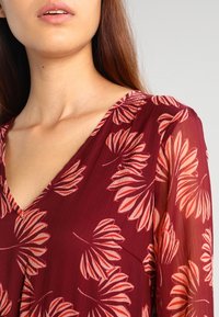 Robe bordeaux avec un décolleté en V, présentant un motif floral dans des tons de rose et de blanc. Fabriquée en tissu léger et transparent.