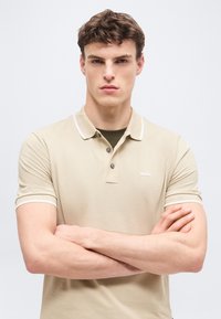 Jeune homme aux bras croisés portant un polo beige BOSS par-dessus un t-shirt foncé, regardant directement la caméra devant un fond uni.