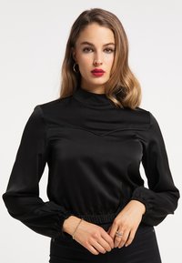 Bluză neagră din satin, cu guler înalt, mâneci pliate și talie ajustată. Prezintă un design subtil în formă de V la nivelul pieptului.