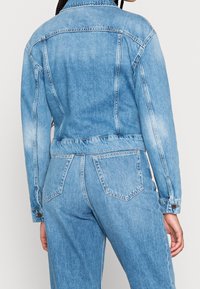 Ljust blå jeansjacka med en figurnära design, synlig sömnad, knappar och två bakfickor. Slät textur med en lätt blekning.