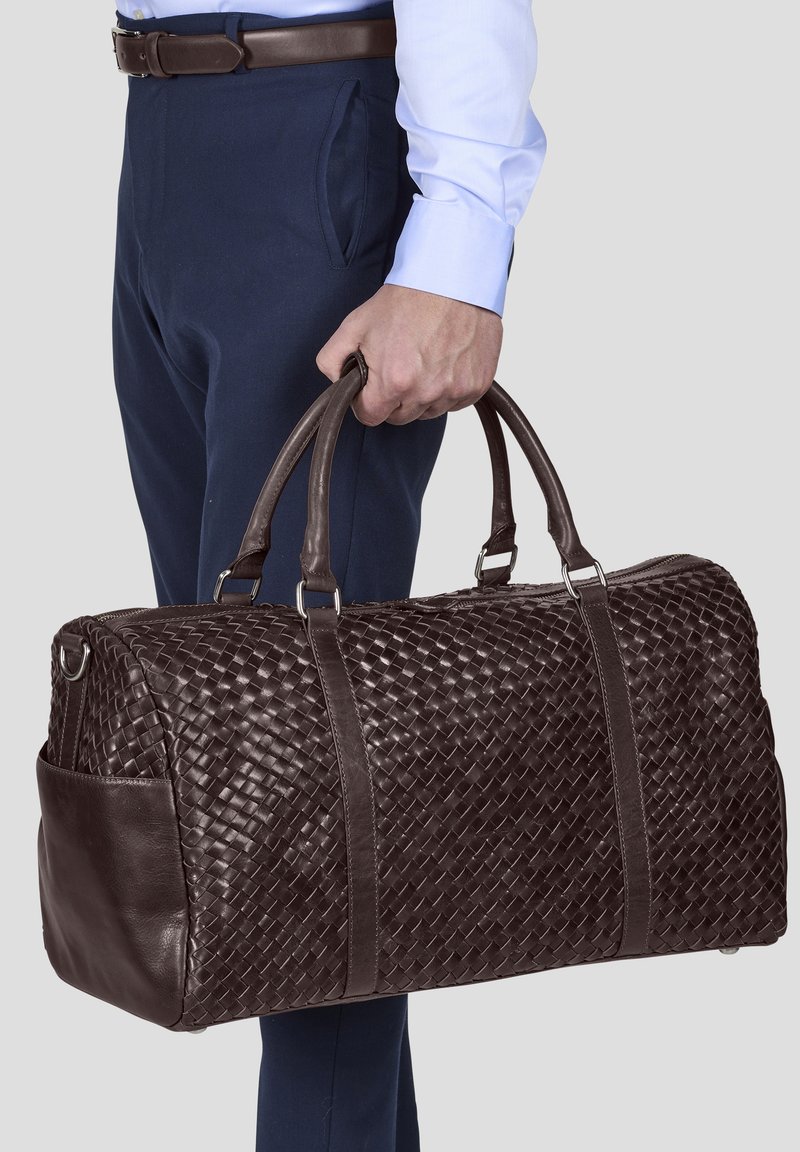 Brun flettet skinn duffelbag med doble håndtak, sølvfarget maskinvare og en sidelomme, presentert mot en nøytral bakgrunn.