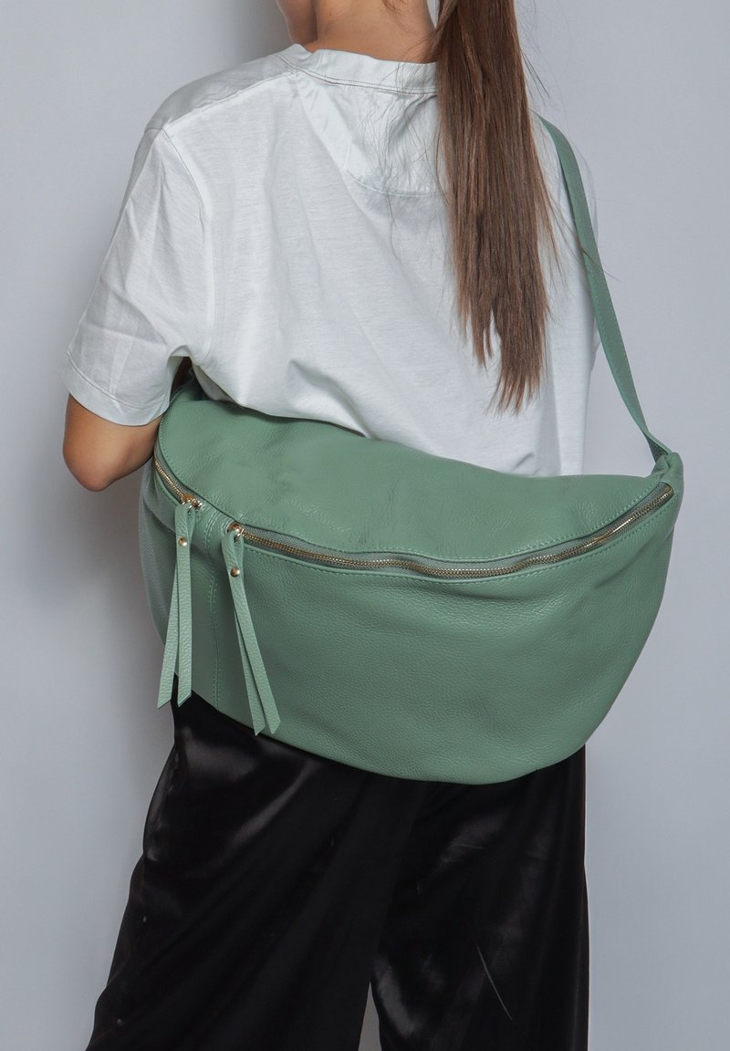 Borsa a tracolla verde in pelle con una superficie liscia, forma curvata, cerniera dorata e doppi dettagli a frange. Indossata sopra una maglietta bianca.