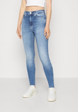 Kvinde iført lyseblå højtaljede skinny jeans med en hvid croppet top og hvide sneakers, stående mod en ensfarvet lys baggrund.