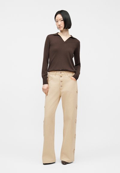 Γυναίκα με κοντά μαλλιά μαύρου χρώματος, φοράει καφέ μπλούζα με μακριά μανίκια και μπεζ παντελόνι ψηλόμεσο με κουμπιά στο πλάι, στέκεται με το ένα χέρι στην τσέπη.