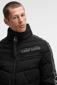 Mann mit kurzen Haaren, der eine schwarze gesteppten Jacke mit dem "CAMP DAVID"-Logo und Reißverschlusstaschen auf der Brust vor schlichtem Hintergrund trägt.