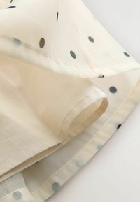 Jasna kremowa tkanina z czarnymi i niebieskimi kropkami, o gładkiej fakturze i widocznym szwie na brzegu oraz wewnętrznej szwie.