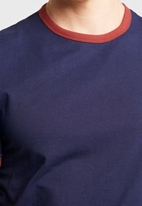 Osoba nosząca granatową koszulkę z okrągłym wycięciem w kolorze rdzy, widoczną od ramion do środka klatki piersiowej.