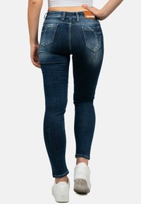 Jeans skinny in denim blu scuro, con vita media, due tasche posteriori e lievi strappi vicino alla vita. Indossati con sneakers bianche.
