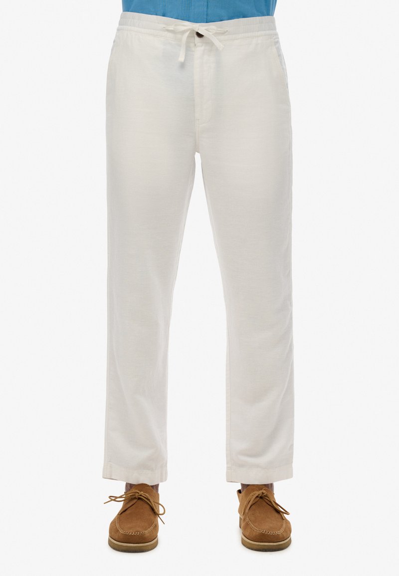 Pantalones de lino blancos con cintura elástica y cordón, corte recto, bajos con puños, combinados con zapatos de ante marrón. Textura sutil visible.