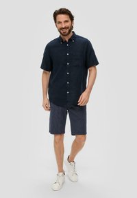 Marine linnen overhemd met korte mouwen en knoopsluiting, gecombineerd met gestreepte marineblauwe shorts. Schoeisel is witte sneakers met veters.