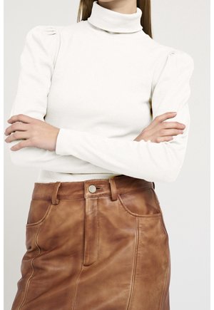 Frau mit verschränkten Armen, die einen weißen Rollkragenpullover und einen braunen Lederrock mit Taschen und Knopfverschluss trägt.