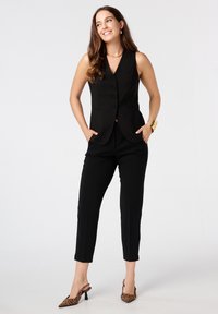 Gilet sans manches noir avec cinq boutons, associé à un pantalon noir ajusté et des talons imprimés animaux. Tissu lisse, design ajusté et accents stylés.
