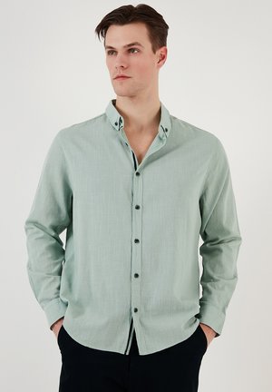 Camisa verde claro de botón en una tela texturizada, con mangas largas, un cuello y botones oscuros, llevada con las manos en los bolsillos.