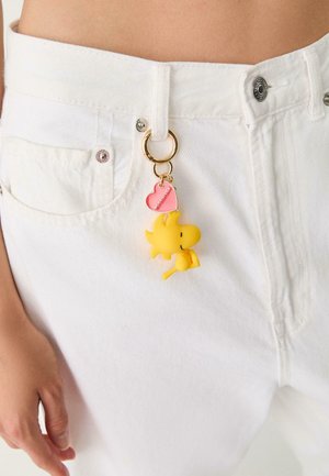 Porte-clés en forme d'oiseau cartoon jaune accroché à un anneau en or, avec une étiquette en forme de cœur rose portant le nom "Woodstock", clipsé à un jean en denim blanc.