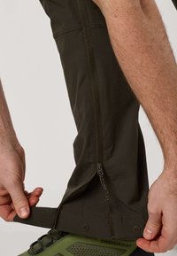 Personne ajustant la fermeture éclair et la sangle au niveau du bas de pantalon sombre de plein air porté par-dessus une chaussure de randonnée verte.