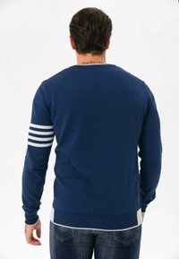 Marineblauer Strickpullover mit langen Ärmeln, der weiße Streifen am linken Arm und einen gerippten Saum aufweist. Glatte Textur und klassischer Schnitt.